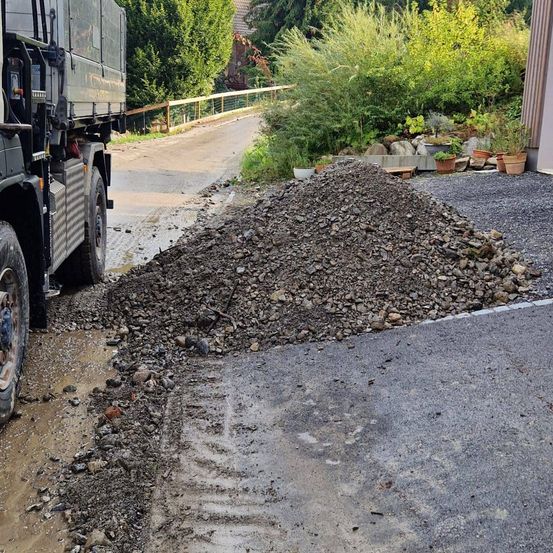 Ein LKW ist auf einer Einfahrt geparkt, wobei Kies auf die Straße verschüttet wurde. Im Hintergrund befinden sich Büsche und Pflanzen sowie ein Holzzaun.