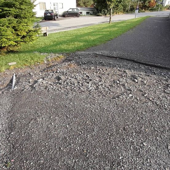 Eine Einfahrt mit einem beschädigten Abschnitt aus Kies und kleinen Steinen neben einem Grasbereich. Zwei Autos sind vor einem weißen Gebäude geparkt.