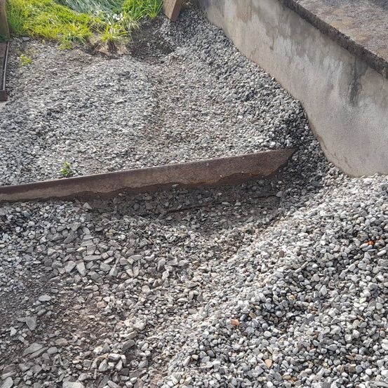 Ein Nahaufnahme einer Betonkante zeigt einen Haufen Kies und kleiner Steine daneben. Ein rostiger Metallstab liegt auf dem Kies.