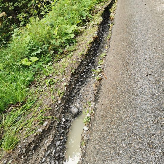 Eine Straße mit einem sichtbaren Schlagloch, teilweise mit Wasser gefüllt, neben einem Grasrand an einem sonnigen Tag.