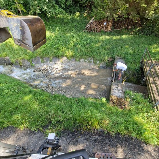 Ein Mann arbeitet an einem Bach, möglicherweise zur Reparatur oder Instandhaltung. Er steht auf Betonstufen in der Nähe eines Haufens von Steinen. In der Nähe befindet sich ein großes Baufahrzeug.