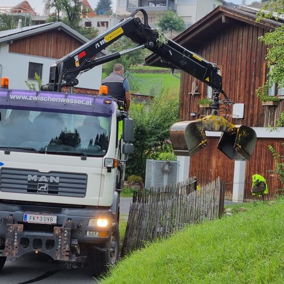 Ein MAN-Lkw mit ausgeklapptem Kran steht am Straßenrand. Eine Person steht im Inneren des Lkw, eine weitere auf dem Kran. Hinter dem Lkw befindet sich ein Holzhaus.