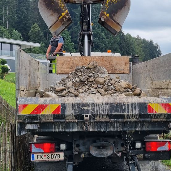 Eine Person bedient einen Kran an einer Baustelle. Der Kran lädt Steine in den Laderaum eines Lastwagens. Der Lastwagen ist am Straßenrand geparkt.