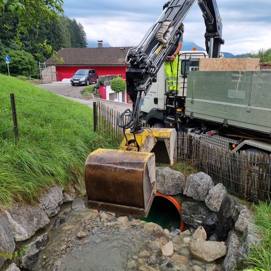 Ein Arbeiter bedient einen großen Bagger, um Steine aus einem Bach zu entfernen, in der Nähe eines roten Hauses mit einem geparkten Auto davor, an einem bewölkten Tag.