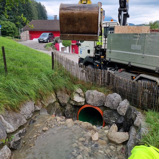 Ein Baufahrzeug hebt eine große rostige Schaufel in der Nähe einer wassergefüllten Grube an, die von Steinen und einem Holzzaun umgeben ist. Hinter dem Zaun sind ein rotes Haus und ein geparktes Auto zu sehen.