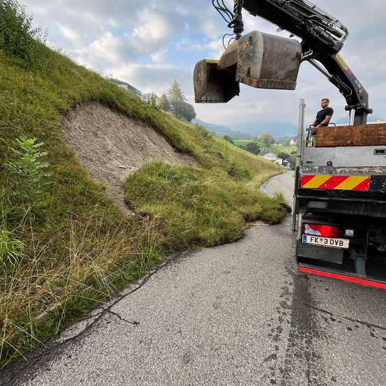 Ein großer Bagger befindet sich auf einem Lastwagen, positioniert über einem Grashang, mit einem Mann im Inneren. Das Kennzeichen des Lastwagens lautet FK 3 DVB.