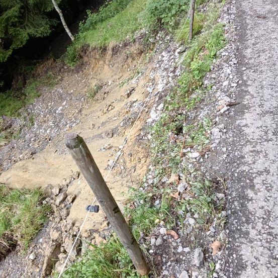 Ein steiles Straßenufer mit Erosion durch Kies und Erde. Ein Holzpfosten und ein Draht sichern den Rand.