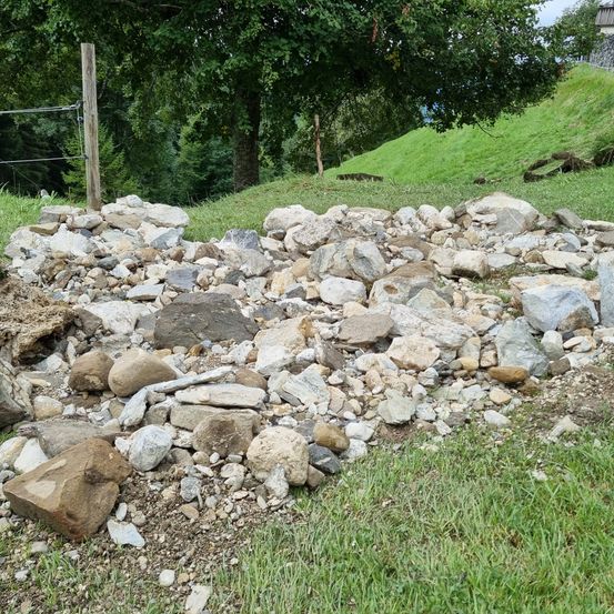 Ein Haufen Steine und Schutt auf einem grasbewachsenen Hügel mit einem Holzpfahl und einem Drahtzaun, umgeben von Bäumen und einem klaren Himmel.