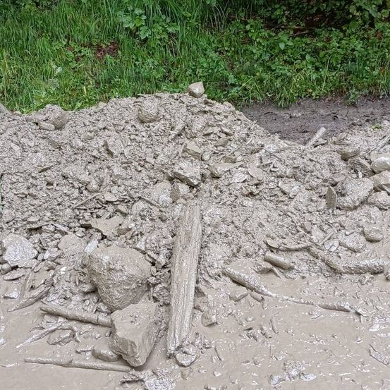 Ein Haufen schlammiger Trümmer mit verstreuten Steinen und Holzstücken auf dem Boden in der Nähe eines Grasbereichs.