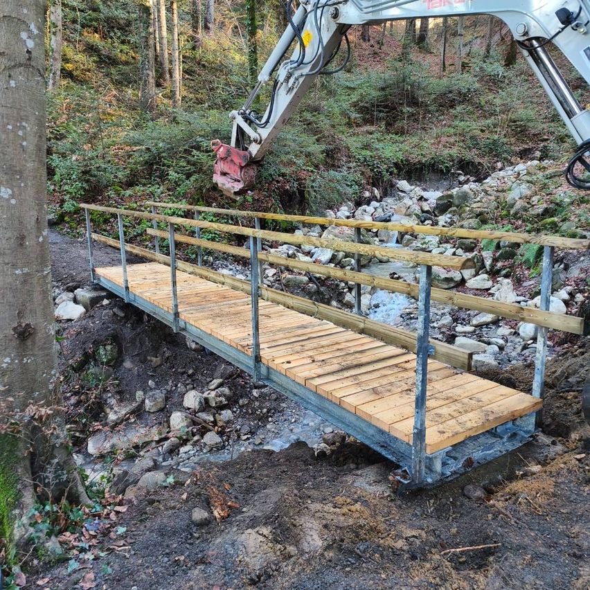 Eine Holzbrücke im Bau in einem Wald, mit einem Metallrahmen, gestützt von einem Baufahrzeug, umgeben von Felsen und Vegetation.