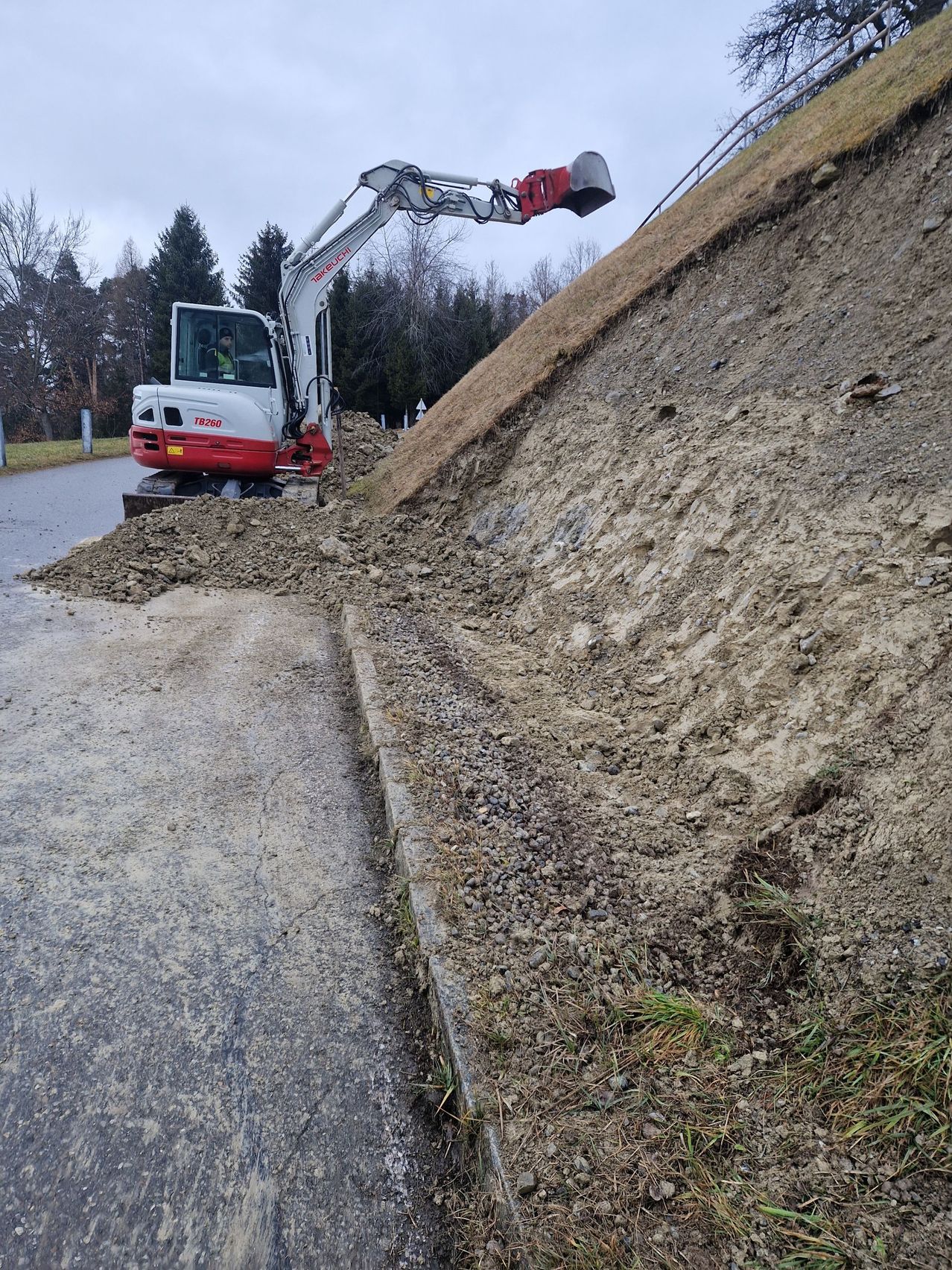 Ein weißer und roter Bagger befindet sich auf einem Hang, mit einer Person im Inneren. Schmutz und kleine Steine sind verstreut. Bäume und Gras befinden sich an der Seite.