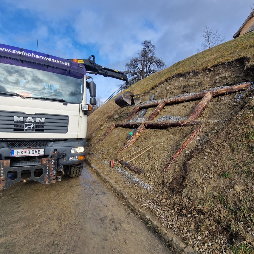 Ein MAN-Lkw mit Kranaufbau steht auf einem Hang mit einer gebrochenen Holzkonstruktion und verstreuten Werkzeugen auf dem Boden.