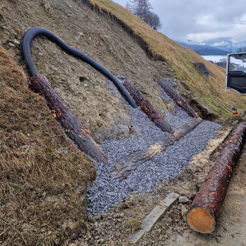 Ein Außenbaustelle zeigt einen großen auf Kies verlegten Schlauch, mit teilweise sichtbaren Baumstämmen und einem Fahrzeug.