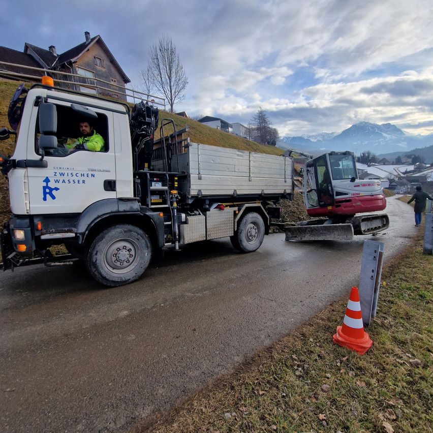 Ein Lkw mit einem Kranaufbau fährt eine Straße hinunter. Der Lkw ist mit Zwischen Wasser beschriftet. Es steht ein Verkehrskegel am Straßenrand. Eine Person steht im Hintergrund.