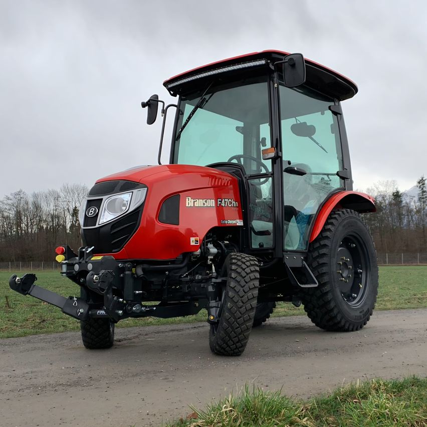 Ein roter Branson F47Chn Traktor mit schwarzem Dach und klarer Windschutzscheibe steht auf einem unbefestigten Weg mit Gras auf beiden Seiten.
