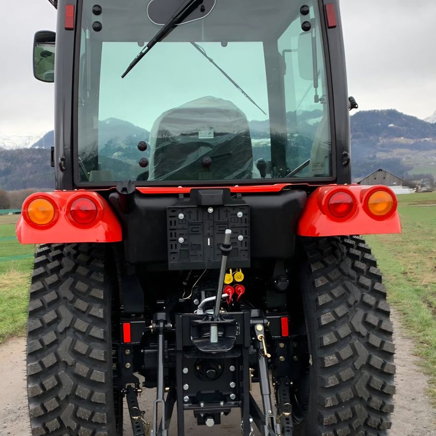 Ein roter Traktor mit schwarzen Reifen steht auf einem Feldweg mit einem Grasfeld und verschneiten Bergen im Hintergrund.
