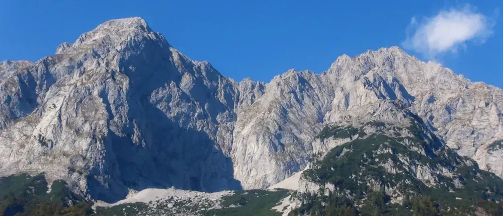 Ein Panoramablick auf majestätische Berge unter einem klaren blauen Himmel. Die rauen Gipfel sind mit Flecken grüner Vegetation und Schnee geschmückt.