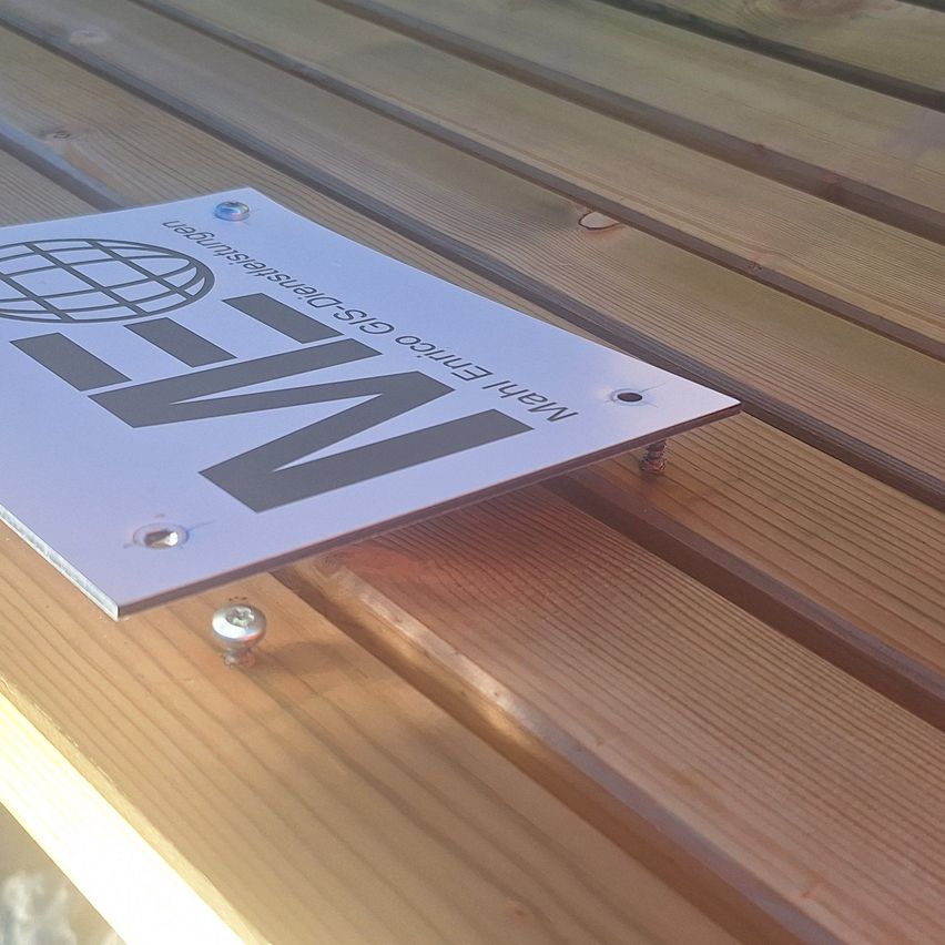 Eine Metallplatte mit 'MW' und einem Globuslogo ist mit Schrauben an einer Holzoberfläche befestigt.