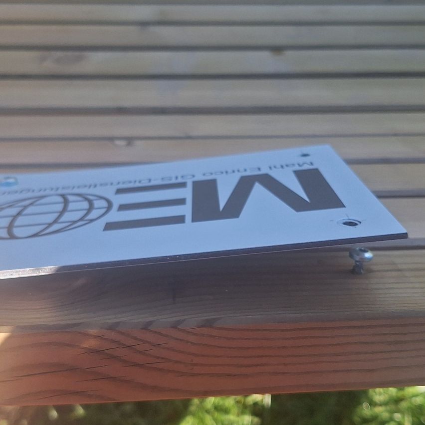 Ein weißes rechteckiges Schild mit dem Wort NEW in großen grauen Buchstaben ist an einer Holzoberfläche befestigt. Das Schild hat einen grauen Rand und ein Globus-Symbol.