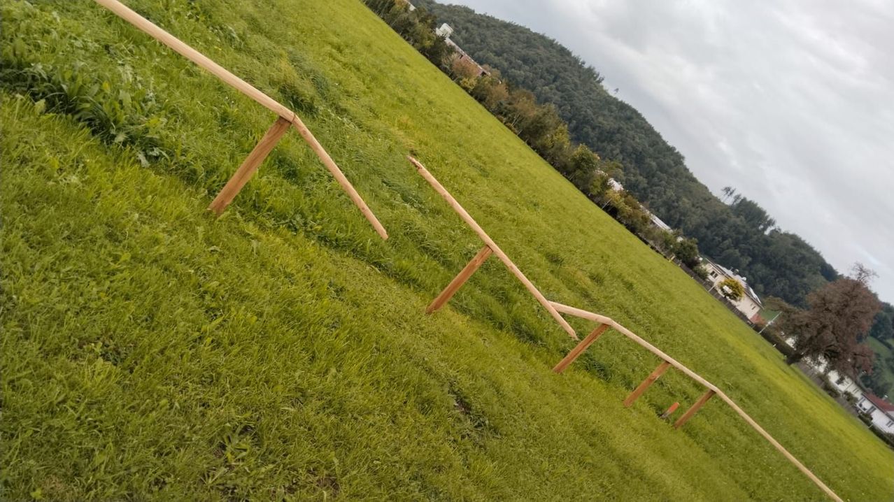 Eine Holzstruktur auf einem Rasenfeld neigt sich nach oben. Der Boden ist mit saftigem grünem Gras bedeckt. Bäume und ein Berg sind in der Ferne sichtbar.
