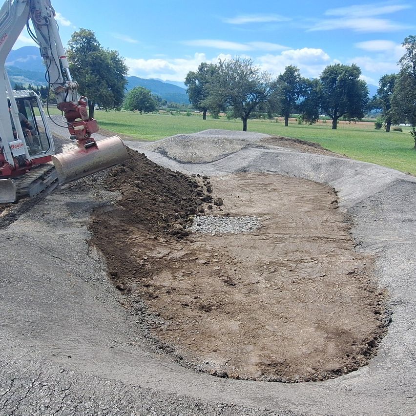 Ein Bagger gräbt ein kreisförmiges Loch auf einer gepflasterten Straße, umgeben von Gras und Bäumen.