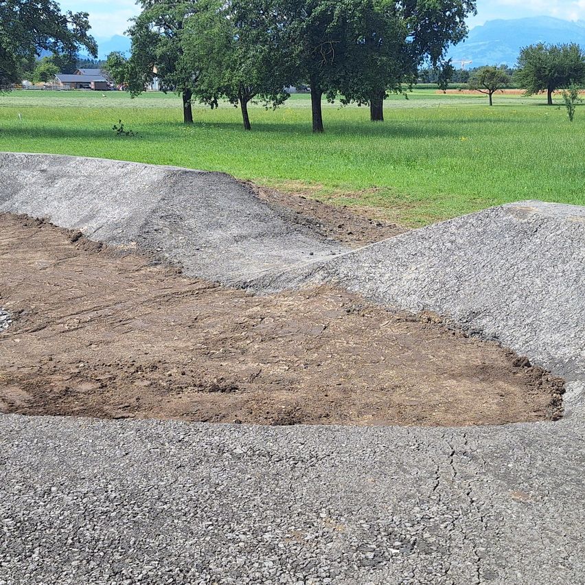 Eine Baustelle mit einer Rampe aus Kies und Erde, umgeben von einem grünen Feld mit Bäumen und einem Gebäude in der Ferne.