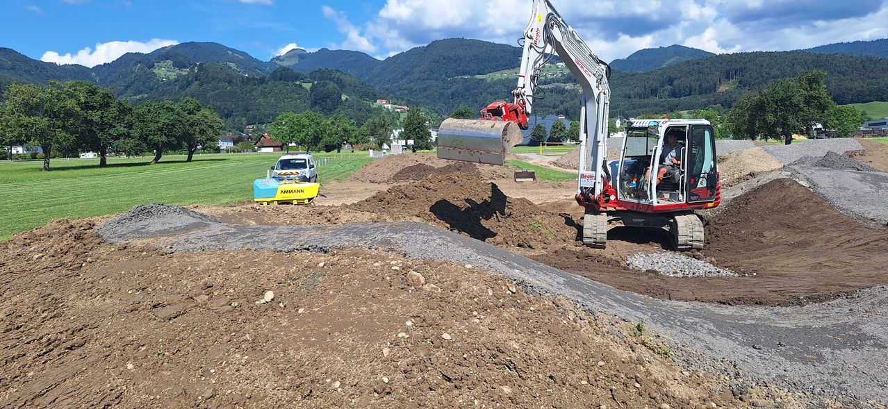 Eine Baustelle mit einem großen Bagger, der Erde ausgräbt, einem geparkten Van in der Nähe und Bergen im Hintergrund.