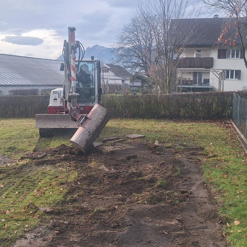 Ein roter und weißer Bagger arbeitet auf einem Grundstück in einem Garten mit einem Haus im Hintergrund.