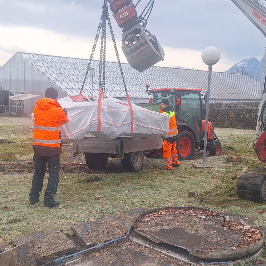 Zwei Arbeiter in orangen Westen benutzen einen Kran, um einen abgedeckten Gegenstand von einem Anhänger zu heben. Ein Traktor steht dahinter mit einer Person im Inneren. Im Hintergrund befindet sich ein Gewächshaus und eine Bergkette.