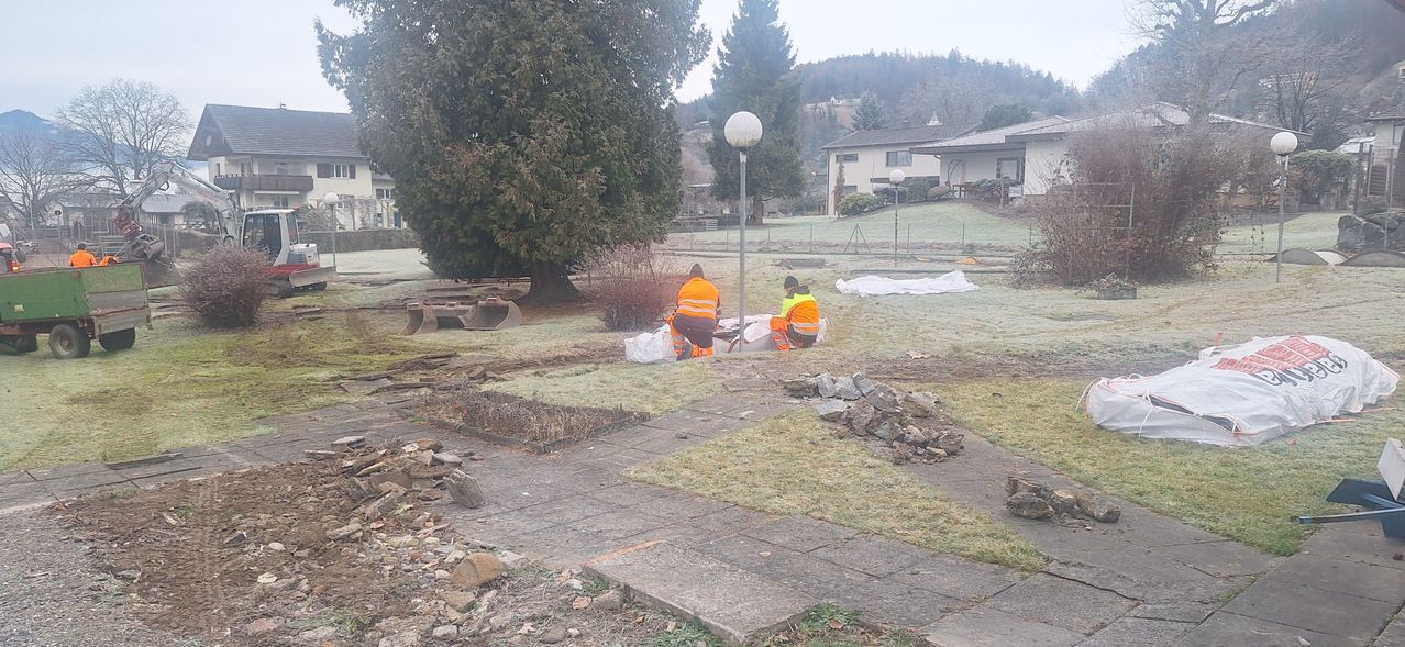 Zwei Arbeiter in orangefarbenen Sicherheitswesten arbeiten an einem Landschaftsprojekt. Sie befinden sich in der Nähe eines Baumes und eines Laternenmastes, mit einem Haufen Steine und etwas Schutt in der Nähe. Ein Gebäude ist im Hintergrund zu sehen.