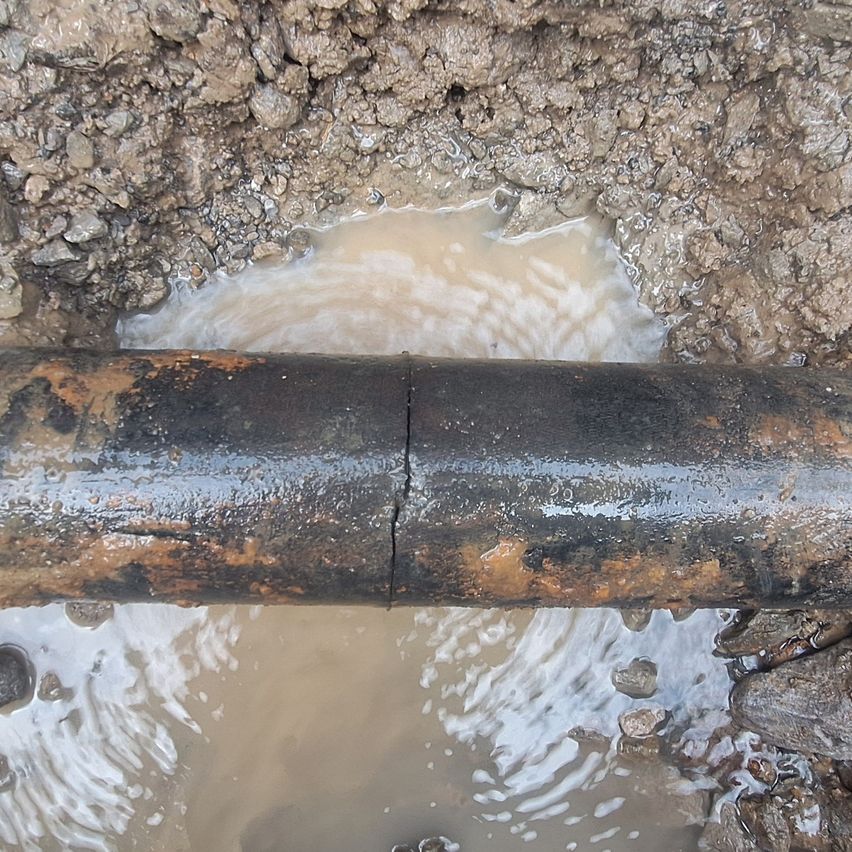 Ein großer, rostiger Metallrohr ist teilweise in schlammiges Wasser eingetaucht, mit sichtbaren Rissen auf seiner Oberfläche.