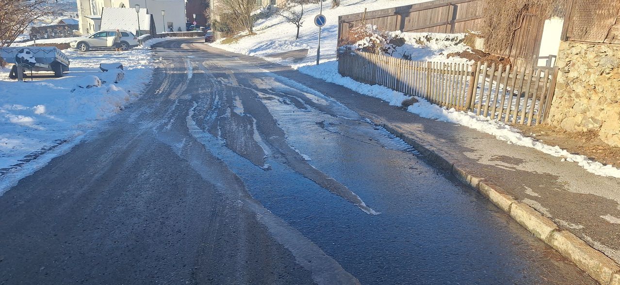 Eine nasse Straße mit Reifenspuren und Pfützen, gesäumt von schneebedeckten Böschungen und Holzzäunen, und einem Verbotsschild.