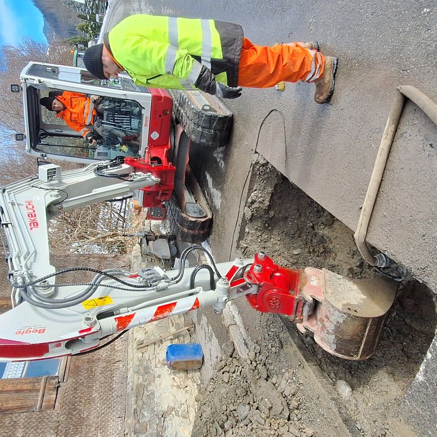 Ein Bauarbeiter in leuchtend orangefarbener Arbeitskleidung benutzt einen rot-weissen Bagger, um in die Strasse zu graben. Ein anderer Arbeiter sitzt in der Fahrerkabine des Baggers.
