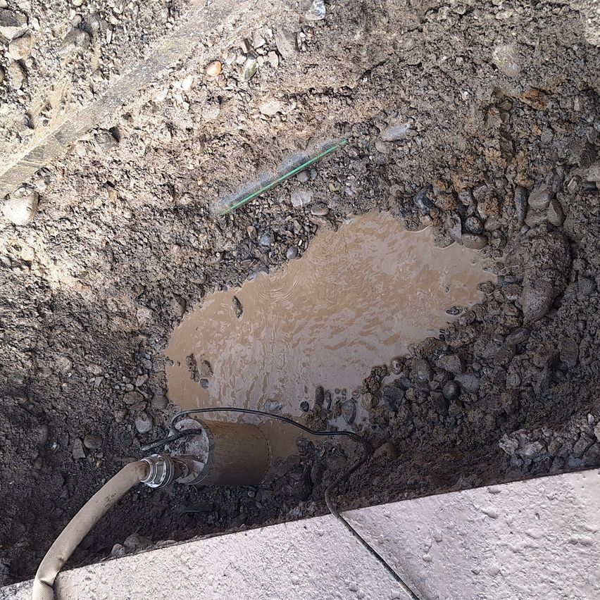 In einem Loch im Boden ist eine schlammige Pfütze sichtbar, mit einem Schlauch und einem Kabel, das an einem Rohr angeschlossen ist.