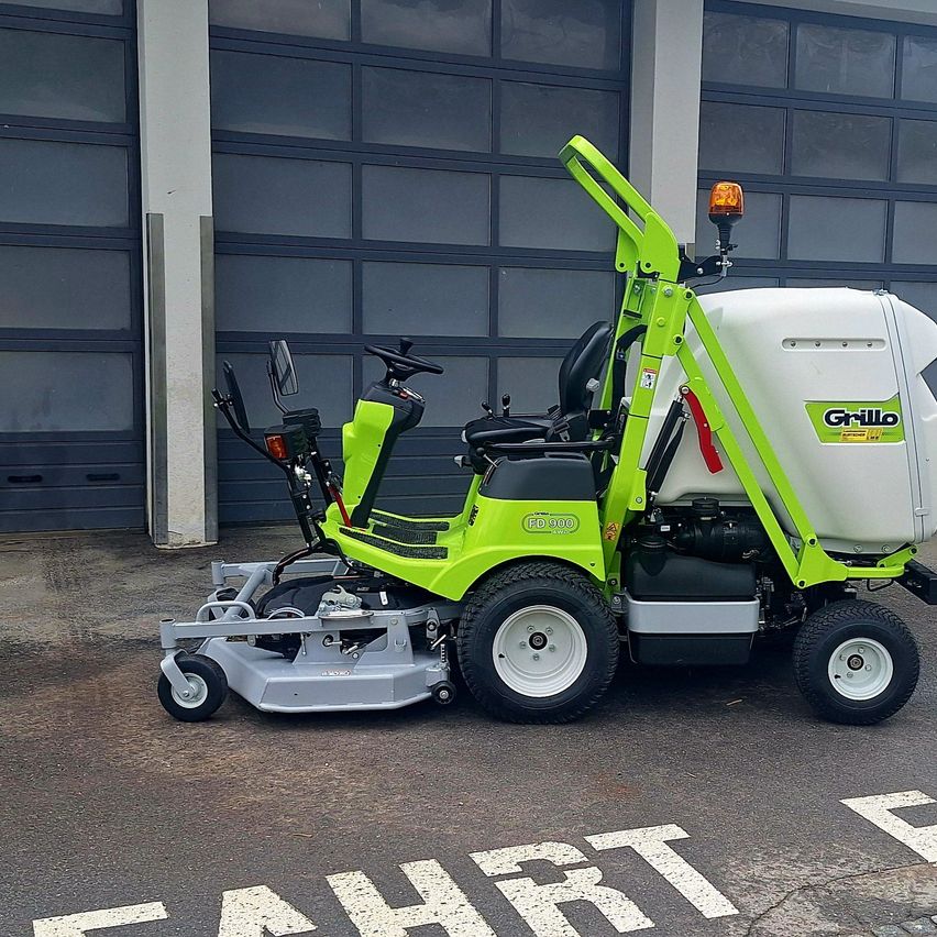 A green and white lawn mower is parked in front of a building with a white "FAHRT" sign on the ground.