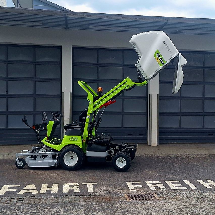A green lawn mower is parked in front of a garage with its lid open. The words 'FAHRT FREIH' are written on the ground.