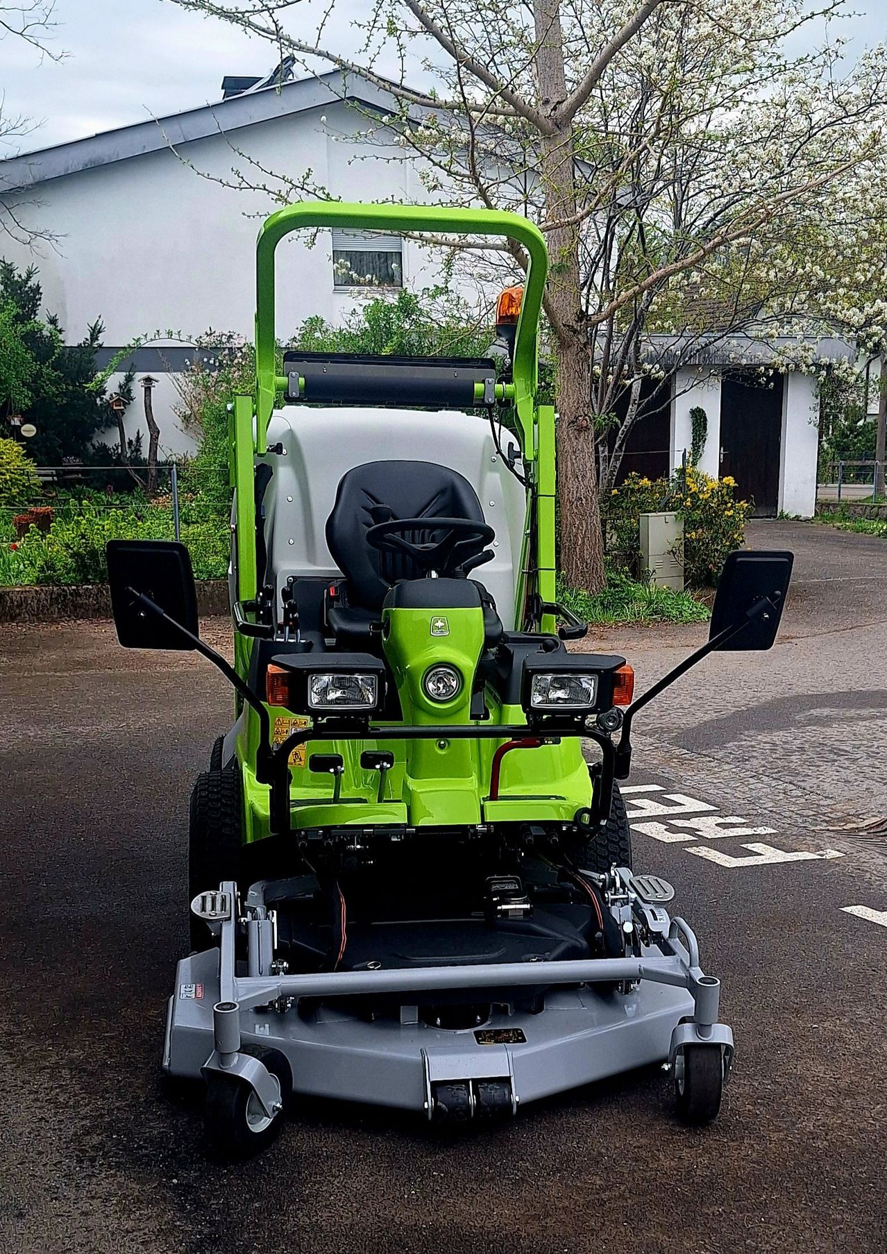 A green and gray tractor is parked on the side of the road with a tree and building in the background.