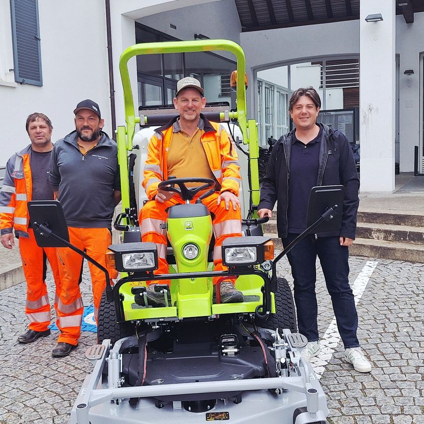 Three men stand in front of a green lawn mower. One man sits in the driver's seat, wearing an orange safety jacket. The other two men stand beside him. Behind them is a white building with glass windows.
