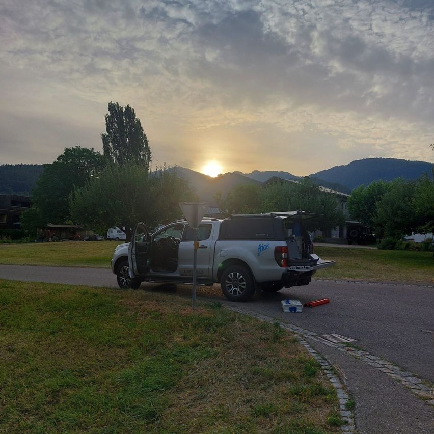A white pickup truck with its back door open is parked on the side of a road with a grassy area and trees. The sun is setting behind the mountains.