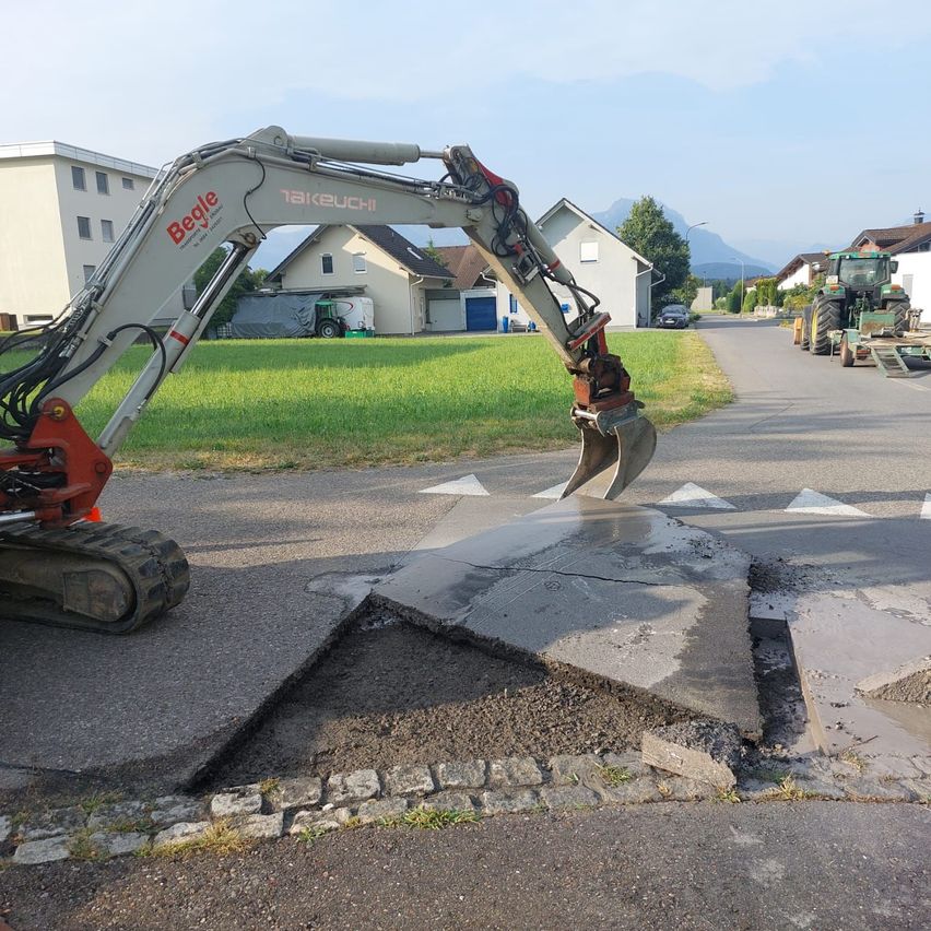 A large construction vehicle with the word 'Begle' on it is lifting a broken section of concrete road. The road is surrounded by houses and a grassy field.