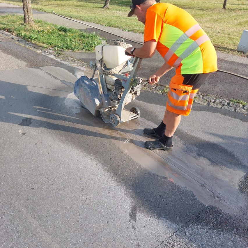 A man in high visibility clothing is using a machine to smooth the road surface. The machine is leaving a wet trail behind it.