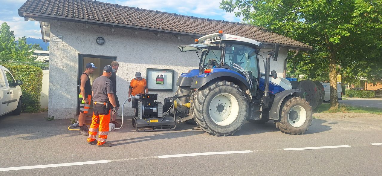 Ein blauer Traktor steht vor einem Gebäude mit einem Schild. Zwei Männer stehen neben dem Traktor, einer hält einen Schlauch.