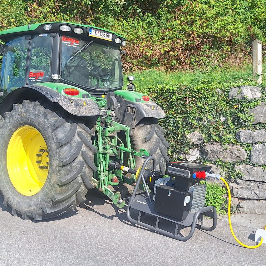Ein grüner Traktor mit einem gelben Rad steht am Straßenrand, angeschlossen an einen Stromerzeuger.