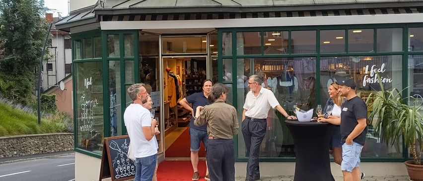 Mehrere Personen versammeln sich vor dem Wiederwertvoll-Laden, einer steht in der Nähe eines Tisches und andere in der Nähe eines Tafels. Ein roter Teppich führt zum Eingang.