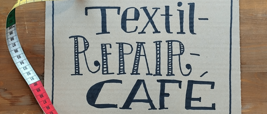 A sign that reads Textil-Repair-Cafe is on a wooden table with a pair of scissors, measuring tape, and spools of thread.