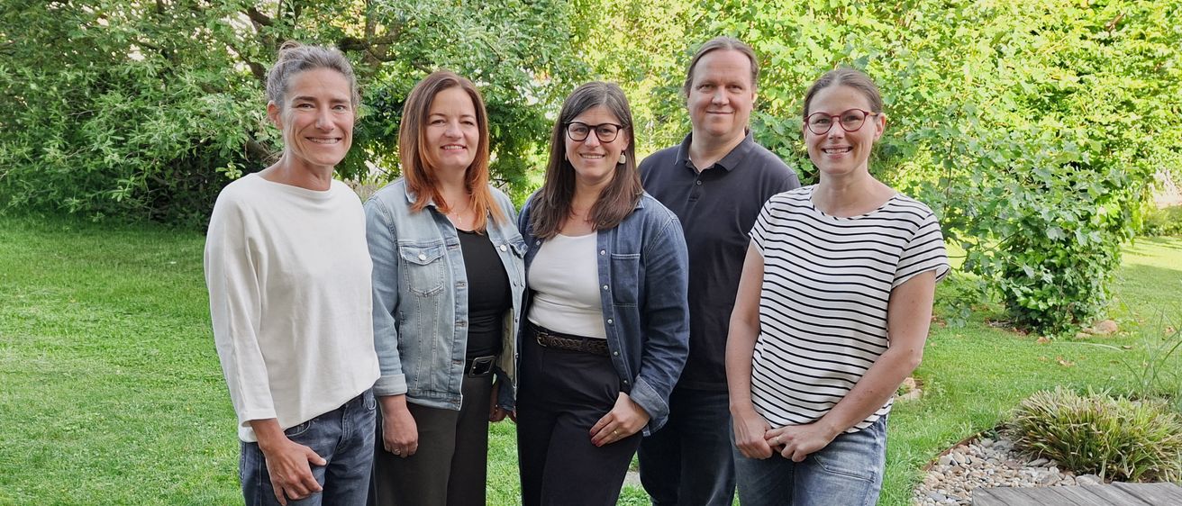 Fünf Personen stehen in einem Garten mit grünem Rasen und Bäumen im Hintergrund. Sie alle lächeln und scheinen für ein Foto zu posieren.