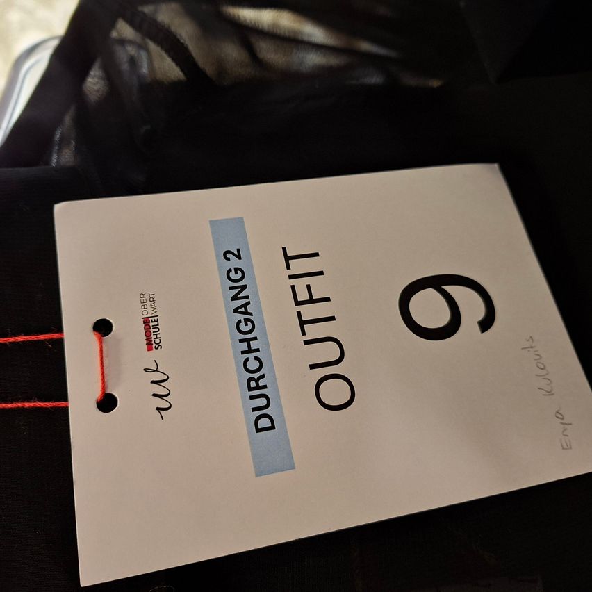 Ein weißer Aufkleber mit dem Text Durchgang 2 und der Nummer 9. Er ist an einem schwarzen Stoff mit einem roten Faden befestigt.