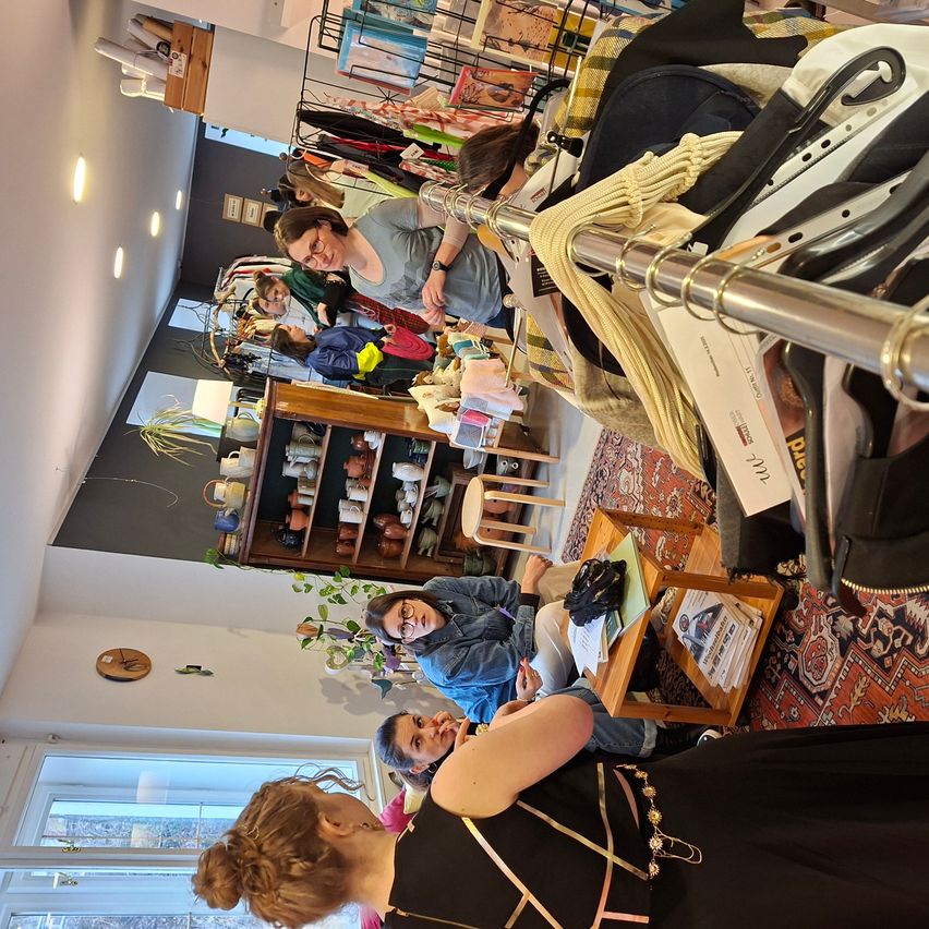 Ein Geschäft mit Kleiderständern, eine Frau mit Brille und Halskette sitzt an einem Tisch mit einer anderen Frau, eine Person sitzt auf einem Stuhl hinter ihnen, ein Holzregal mit Teekannen und Tassen und ein Teppich auf dem Boden.