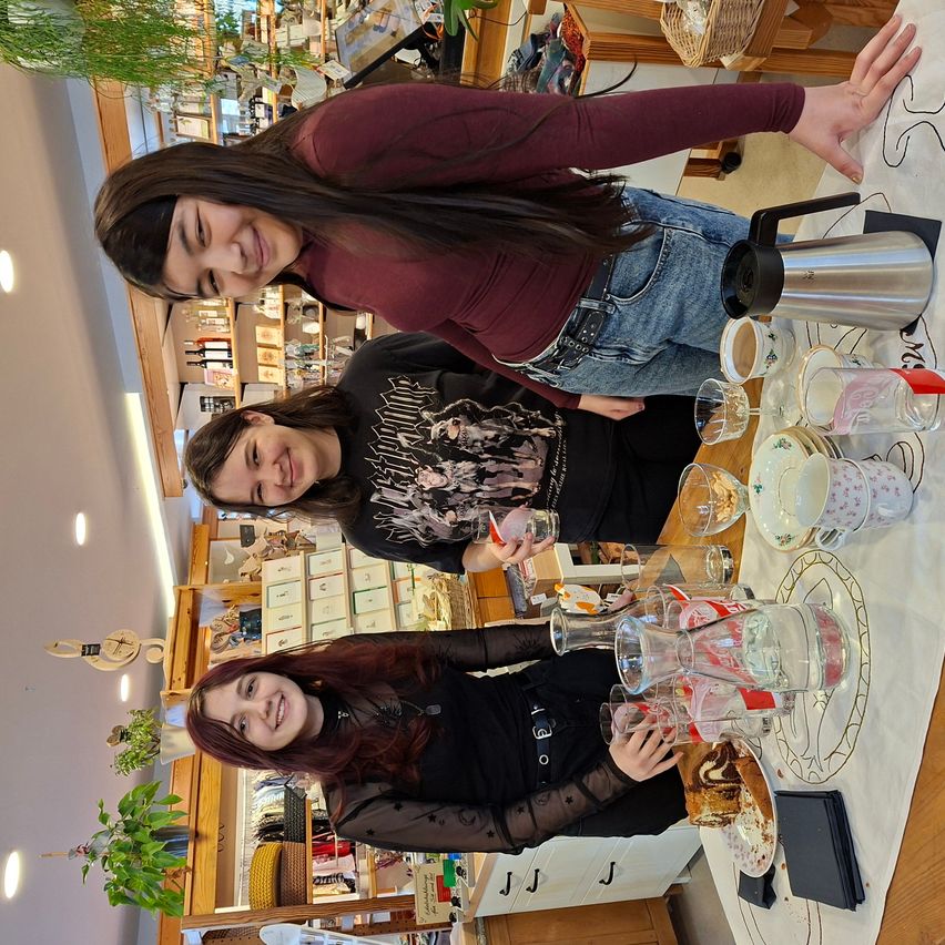 Drei Frauen lächeln an einem Tisch mit Teetassen, Tellern und einem silbernen Kaffeetopf. Eine Frau in einem weinroten Shirt lehnt sich an den Tisch. Der Hintergrund zeigt ein Geschäft mit verschiedenen Artikeln in den Regalen.