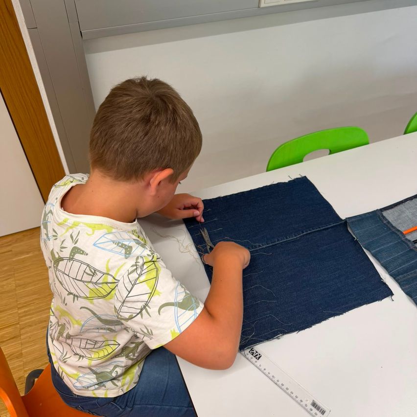 A young boy is seated at a table, using a needle and thread to sew a piece of denim fabric. He is wearing a dinosaur-themed shirt.